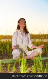 Beautiful young woman doing yoga sitting near the lake early in the morning