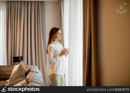 Beautiful young woman drinking coffee and looking through window while standing in the contemporary apartment