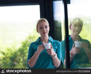 beautiful young woman drinking morning coffee by the window in her home