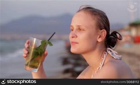 Beautiful young woman enjoying an evening cocktail sitting overlooking a tropical beach with her face turned towards the setting sun