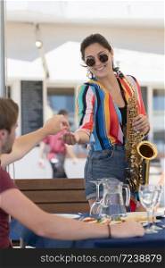 Beautiful young woman holding a saxophone smiling while receiving a tip from a young man sitting at an outdoor bar terrace on an out of focus background. Street performance and lifestyle concept.. woman holding a saxophone smiling while receiving a tip from a young man sitting at an outdoor bar terrace