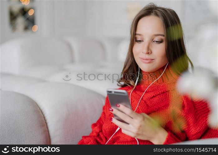 Beautiful young woman holds cell phone, listens music from playlist, surfes social networks and messages with friends, enjoys calm atmosphere at home, sits on comfortable sofa. Lifestyle concept