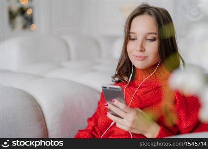 Beautiful young woman holds cell phone, listens music from playlist, surfes social networks and messages with friends, enjoys calm atmosphere at home, sits on comfortable sofa. Lifestyle concept