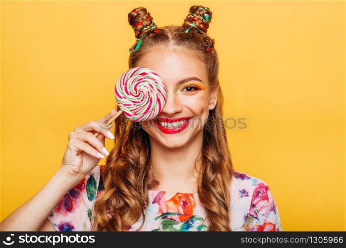 Beautiful young woman holds in hand candy, emotion of indignation on her face. Stylish girl in summer colorful dress. Studio portrait, yellow wall on background.. Beautiful young woman holds in hand candy