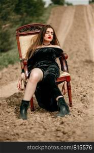 Beautiful young woman in a black dress poses for a photographer sitting on a classic armchair on a sandy road