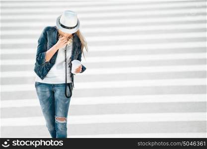 Beautiful young woman in a hat and jeans jacket with a takeaway coffee cup, standing on the road with zebra crossing, drinking coffee, and posing against road background.
