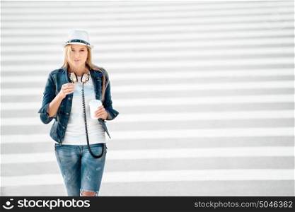 Beautiful young woman in a hat and jeans jacket with a takeaway coffee cup, standing on the road with zebra crossing, drinking coffee, and posing against road background.