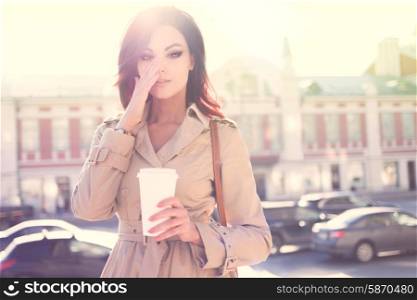 Beautiful young woman in a modern trench coat, holding a disposable takeaway cup and standing against urban city background.