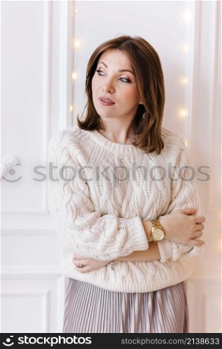 beautiful young woman in a white sweater, skirt and high heels on a background of white wall and christmas garlands.. beautiful young woman in a white sweater, skirt and high heels on a background of white wall and christmas garlands