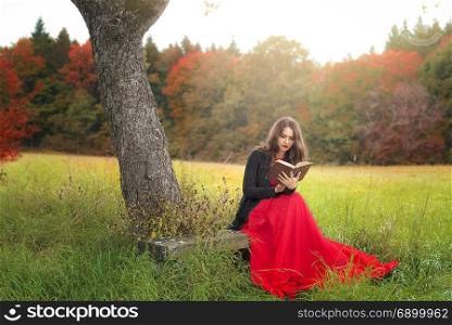 Beautiful young woman, in an elegant red dress and jacket, sitting on a wooden bench, under an old tree, reading an antique book, in a colorful autumn decor.