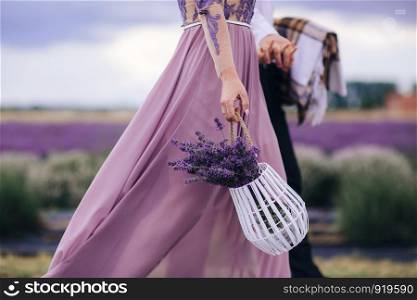 Beautiful young woman in blue dress holds bouquet of flowers lavender in basket while walking outdoor through wheat field at sunset in summer. Provence, France. Toned image with copy space. Beautiful woman holds bouquet of flowers lavender in basket while walking outdoor through wheat field in summer.