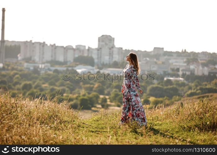 beautiful young woman in dress in floral print walking in the field at sunset. stylish romantic girl with long hair have a good time outdoors on city background. copy space. beautiful young woman in dress in floral print walking in the field at sunset. stylish romantic girl with long hair have a good time outdoors on city background. copy space.