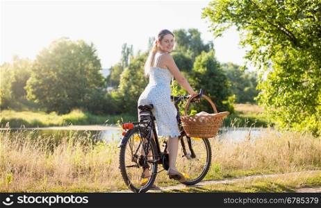 Beautiful young woman in dress riding a bicycle by the river at hot sunny day