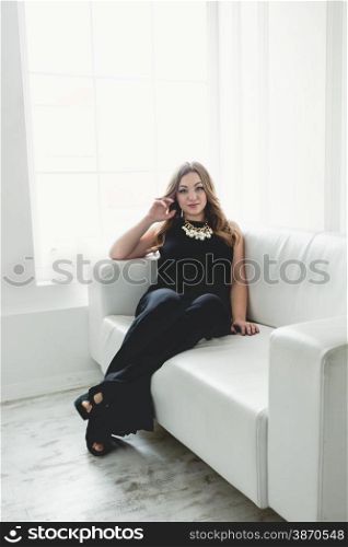 Beautiful young woman in long dress relaxing on white couch next to window