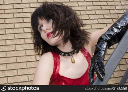Beautiful young woman in red and black vinyl outfit leaning on iron staircase outdoors.