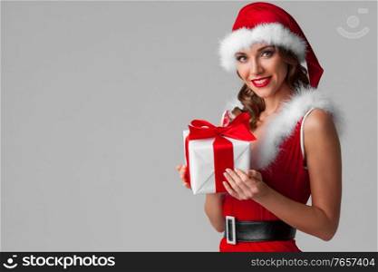 Beautiful young woman in red Santa Claus style clothes holding Christmas gift, gray background. Santa girl with Christmas gift
