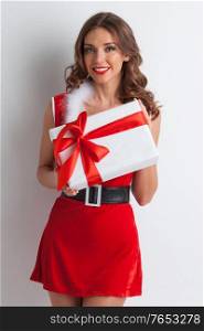 Beautiful young woman in red Santa Claus style clothes holding Christmas gift, white background. Santa girl with Christmas gift