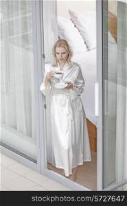 Beautiful young woman in robe having coffee at balcony doorway