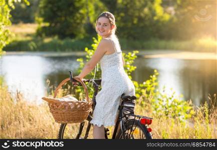 Beautiful young woman in short dress riding on bicycle by the lake at sunset