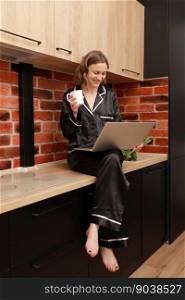 Beautiful young woman in stylish pyjama drinking coffee or tea during her breakfast and working with laptop at home. Smiling girl sits on countertop in kitchen. SMM manager enjoying working at home. Beautiful young woman in stylish pyjama drinking coffee or tea during her breakfast and working with laptop at home. Smiling girl sits on countertop in kitchen. SMM manager enjoying working at home.