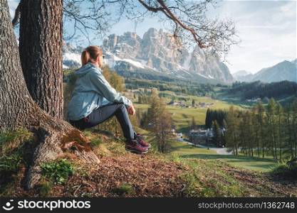 Beautiful young woman in sunglasses and blue jacket is sitting on the hill under the tree at sunset. Colorful spring landscape with sporty girl, meadows, green trees and mountains. Travel in Italy