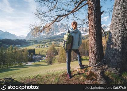 Beautiful young woman in sunglasses and blue jacket is standing on the hill under the tree at sunset. Colorful spring landscape with sporty girl, meadows, green trees and mountains. Travel in Italy