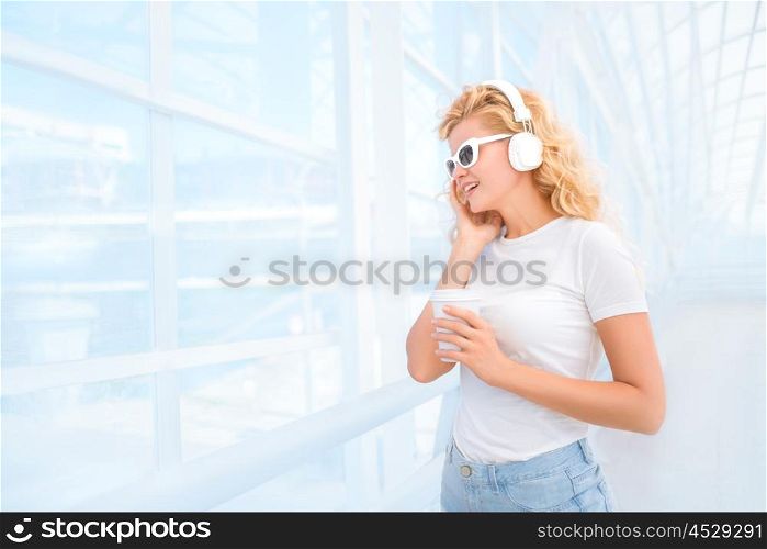 Beautiful young woman in sunglasses with music headphones, standing on the bridge with a take away coffee cup and smiling.