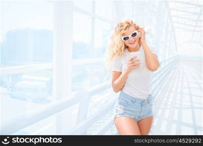 Beautiful young woman in sunglasses with music headphones, standing on the bridge with a take away coffee cup and smiling.