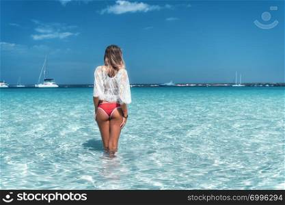 Beautiful young woman in white lace dress in transparent sea at sunny bright day in summer. Tropical landscape. Sexy back of slim girl, clear water, yachts, sky. Tanned woman on the beach. Travel