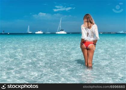 Beautiful young woman in white lace dress in transparent sea at sunny bright morning in summer. Tropical landscape. Sexy back of slim girl, clear water, yachts, sky. Tanned woman on the beach. Travel