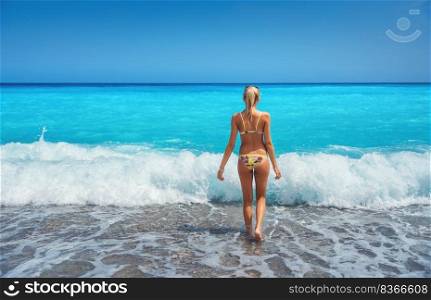 Beautiful young woman in yellow bikini walking in blue sea with waves in bright summer day. Sexy back of girl at the sea coast. Tanned woman relaxing on the sandy beach. Ocean with turquoise water. Beautiful young woman in yellow bikini standing in blue sea