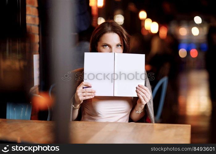 beautiful young woman is holding and covering her face of book with mockup in cafe. Business and studying concept.. beautiful young woman is holding and covering her face of book with mockup in cafe. Business and studying concept