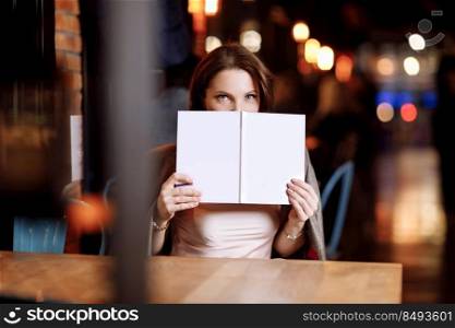 beautiful young woman is holding and covering her face of book with mockup in cafe. Business and studying concept.. beautiful young woman is holding and covering her face of book with mockup in cafe. Business and studying concept