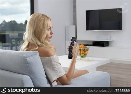 Beautiful, young woman is sitting on the blue sofa in front of tv. She wears comfortable jumper she is looking at camera.