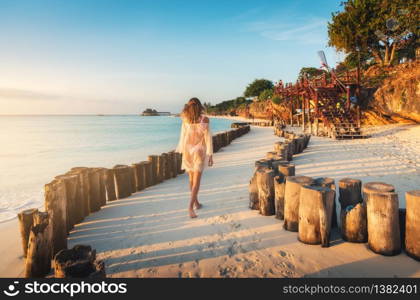 Beautiful young woman is walking on the sandy beach near blue sea at sunset. Summer travel. Tropical landscape with slim girl in white lace dress on the sea coast, wooden stumps, blue sky. Vacation. Beautiful young woman is walking on the sandy beach near blue se