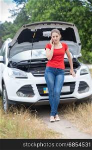 Beautiful young woman leaning on broken car and calling auto service