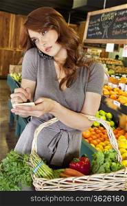 Beautiful young woman listening to mobile phone while making a note of shopping list in market