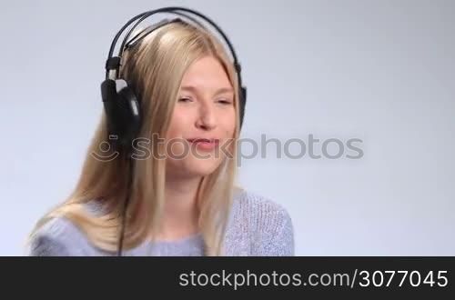 Beautiful young woman listening to music in black stylish headphones on white background. Attractive girl in headphones subtly swaying along with the music, gazing at the camera a little bit shy and starting to flirt with cam.