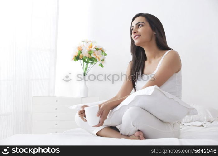 Beautiful young woman looking away while holding coffee cup in bedroom