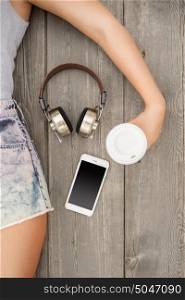 Beautiful young woman lying on the wooden floor with music headphones, smartphone and a take away coffee cup; view from above.