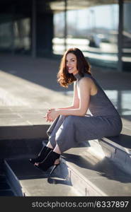 Beautiful young woman, model of fashion, sitting in urban background. Girl smiling.