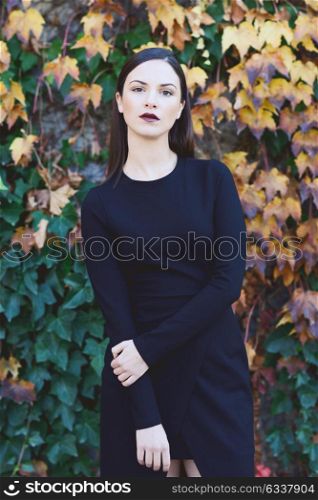 Beautiful young woman, model of fashion, wearing black dress in urban background with autumn colors. Very straight hair styling