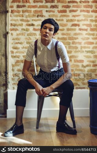 Beautiful young woman, model of fashion, with very short haircut. Beautiful young woman, model of fashion, with very short haircut, looking at camera sitting on chair, with a brick wall in the background. Beauty and hairstyles concept.