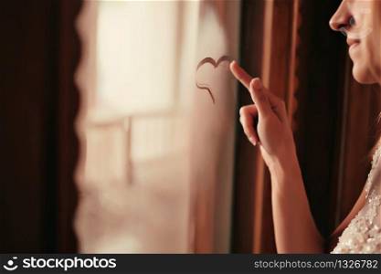 beautiful young woman near the window, draws on glass heart symbol of love. bride&rsquo;s morning.. beautiful young woman near the window, draws on glass heart symbol of love. bride&rsquo;s morning