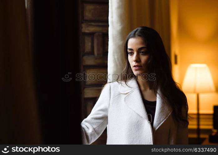 Beautiful young woman posing near a window in her bedroom. Brunette girl wearing white coat.