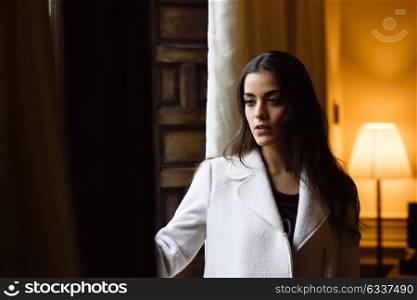 Beautiful young woman posing near a window in her bedroom. Brunette girl wearing white coat.