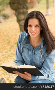 Beautiful young woman reading a book sitting on a bench in park at fall