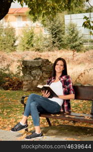 Beautiful young woman reading a book sitting on a bench in park at fall