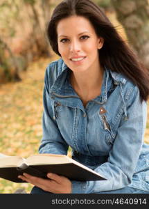 Beautiful young woman reading a book sitting on a bench in park at fall