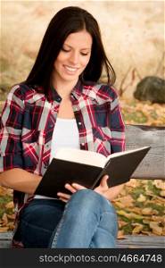 Beautiful young woman reading a book sitting on a bench in park at fall